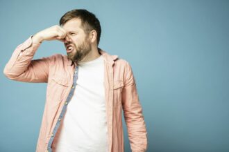 Bearded man squeezes nose with fingers because of an unpleasant odor. Isolated on a blue background.